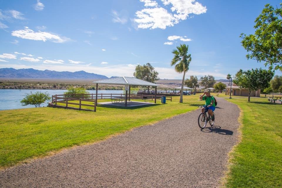 Cyclist riding along the scenic riverside trail at Bullhead City Community Park, with shaded picnic pavilions, palm trees, and distant mountains under a clear blue sky.