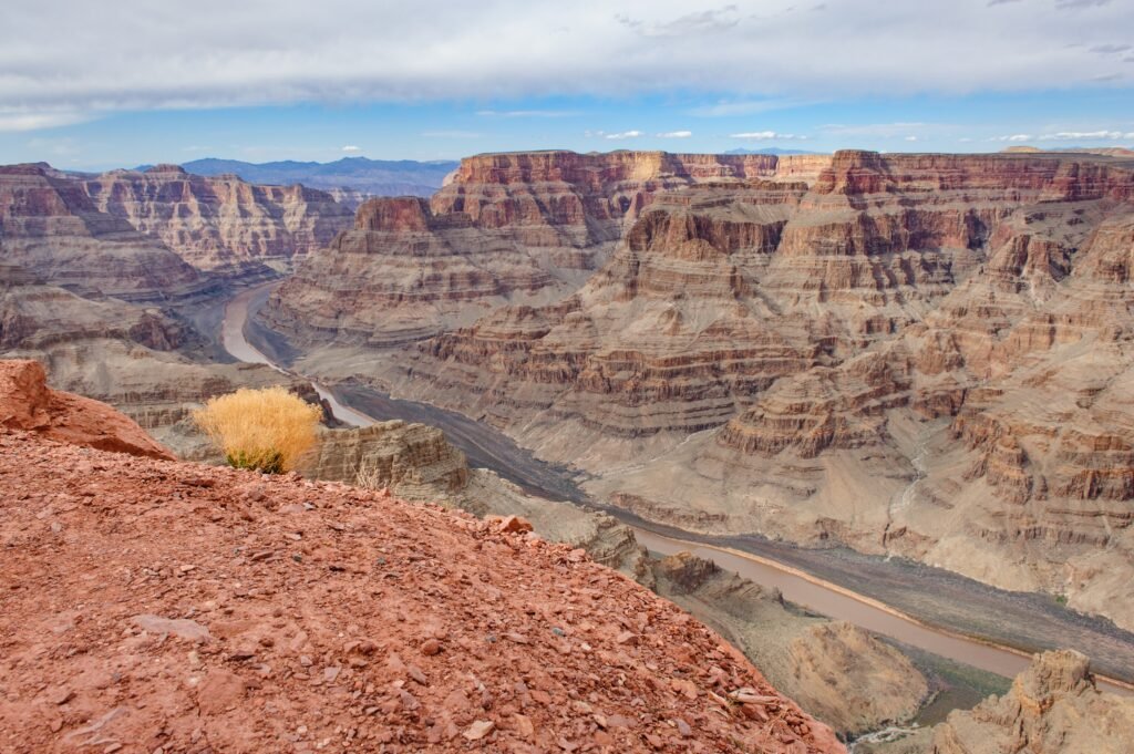 Panoramic view of the Grand Canyon from a rocky overlook, featuring layered red-rock cliffs and the Colorado River winding through the canyon under a cloudy sky.