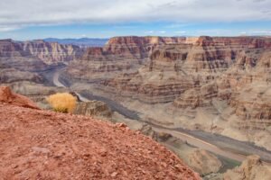 Panoramic view of the Grand Canyon from a rocky overlook, featuring layered red-rock cliffs and the Colorado River winding through the canyon under a cloudy sky.