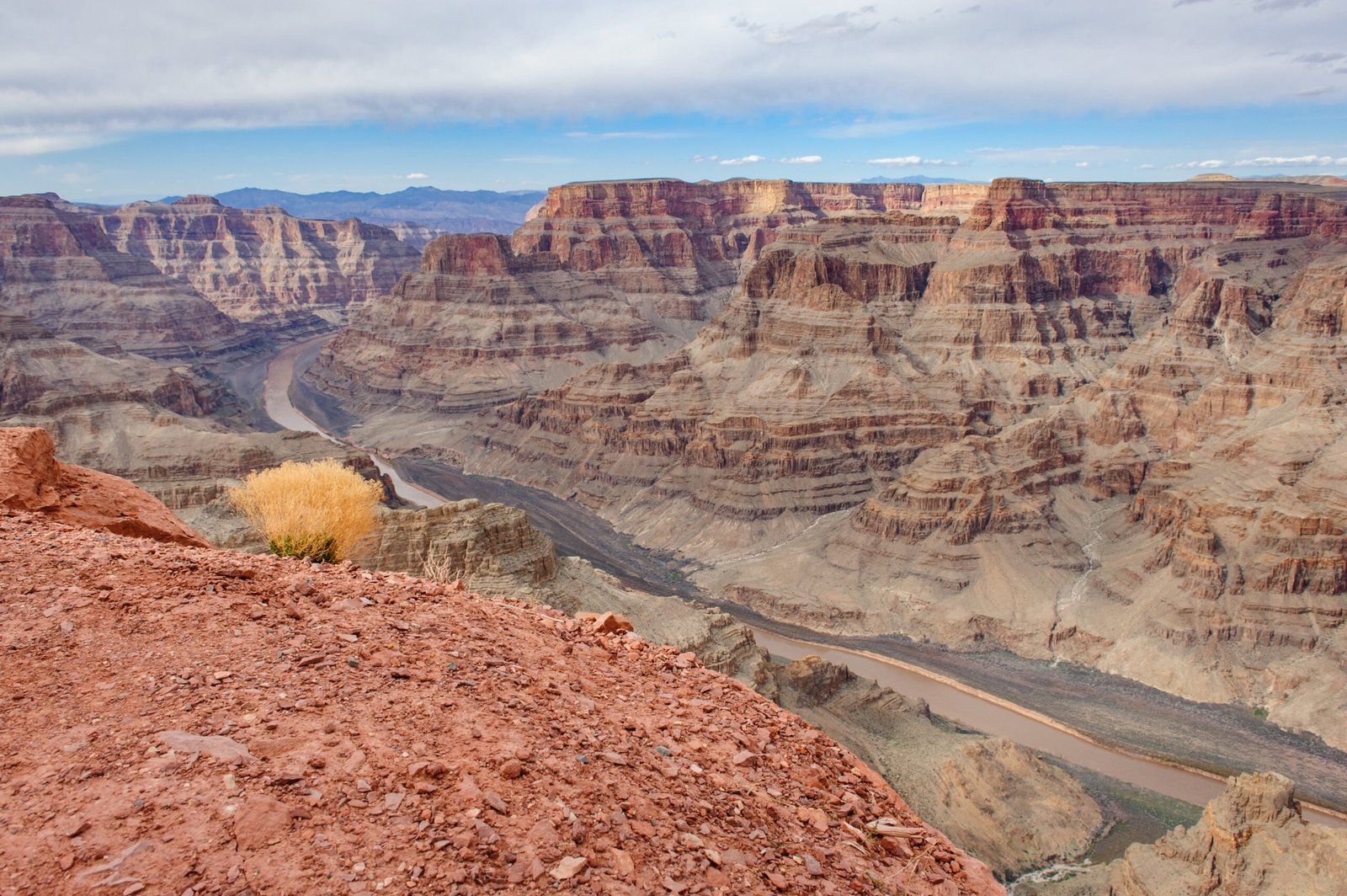 Panoramic view of the Grand Canyon from a rocky overlook, featuring layered red-rock cliffs and the Colorado River winding through the canyon under a cloudy sky.