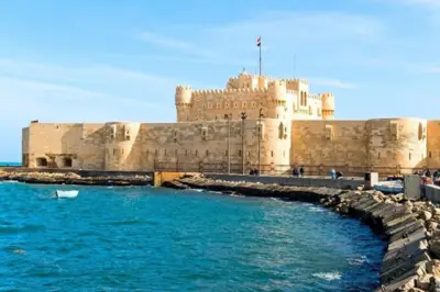 A historic stone fortress overlooking the blue Mediterranean Sea, with a flag flying atop its central tower. The fortress sits at the edge of a curved stone jetty, with a clear blue sky above.