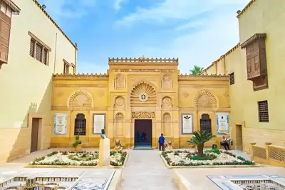 A stunning view of the Coptic Museum entrance in Cairo, Egypt, showcasing intricate Islamic and Coptic architectural details in warm sandstone tones. The courtyard features manicured gardens, statues, and visitors enjoying the cultural site under a bright blue sky.