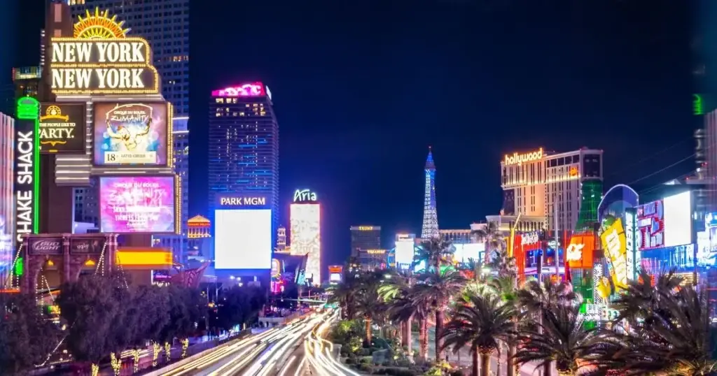 Nighttime view of the vibrant Las Vegas Strip, showcasing famous landmarks such as the New York-New York Hotel, Park MGM, and Aria Resort, illuminated by colorful neon lights and bustling with activity.