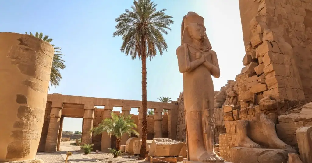 Surrounded by towering columns and palm trees, the colossal statue stands tall amidst the ancient ruins, representing the grandeur and architectural brilliance of ancient Egyptian civilization. The bright sunlight highlights the weathered stone of the temple and statue, offering a glimpse into the rich history of Egypt's New Kingdom period.